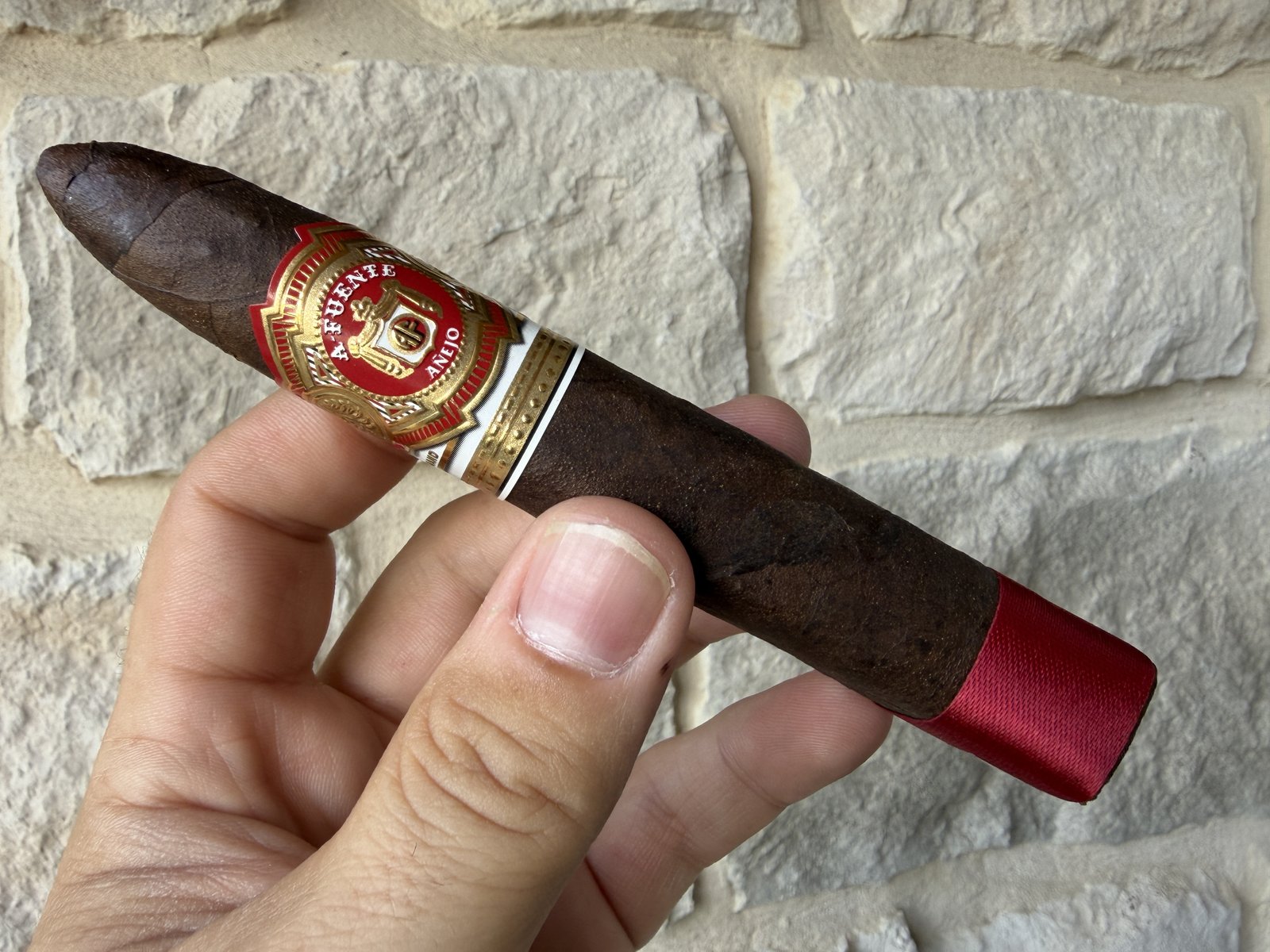 Unlit Arturo Fuente Añejo No. 77 Shark held in hand against a stone wall, dark rustic maduro wrapper, red and gold Añejo band, red satin ribbon protecting the foot