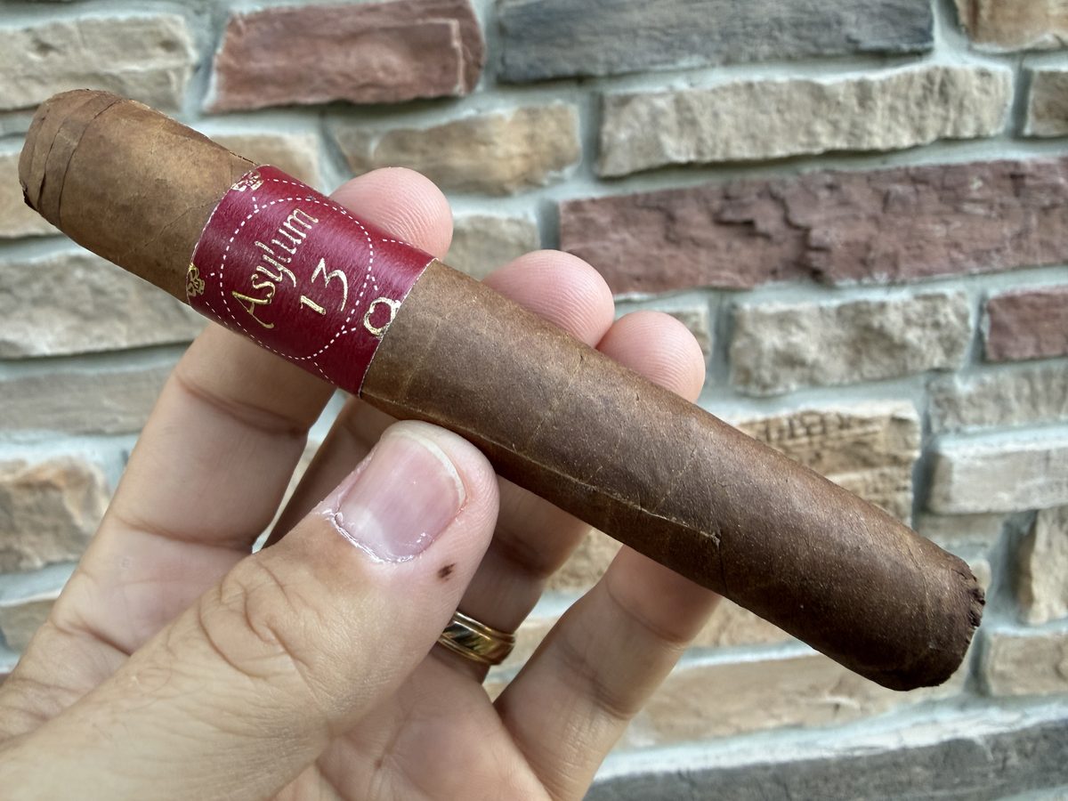 Asylum 13 Corojo Robusto held in hand before lighting, clean veiny medium-brown wrapper with the red Asylum 13 band