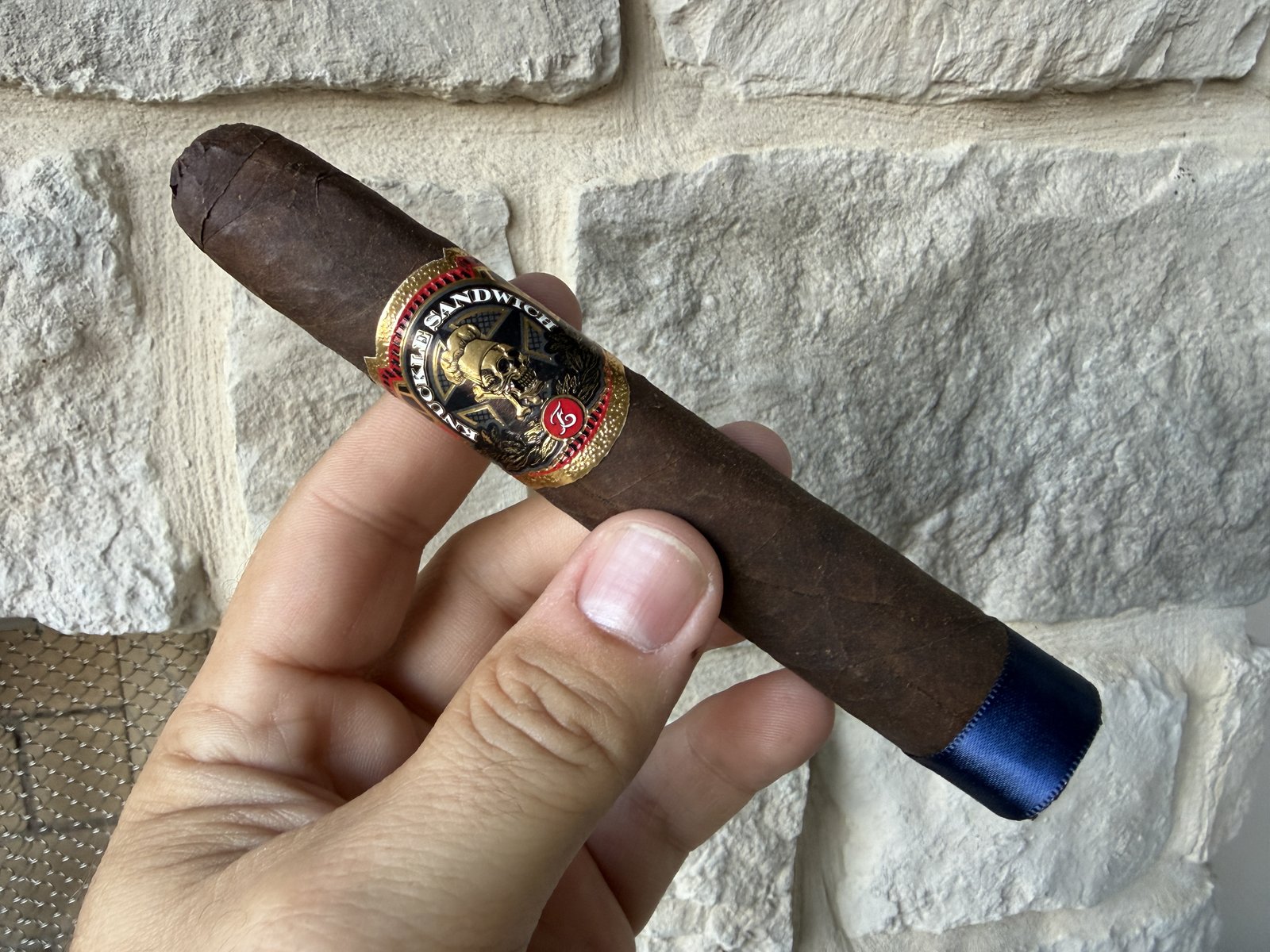 Unlit Espinosa Knuckle Sandwich Chef Special held over a stone wall, dark brown Connecticut Broadleaf wrapper with the black-and-gold skull band and a blue satin ribbon still on the foot