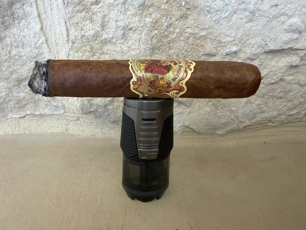 A lit My Father Flor de las Antillas Toro resting across the top of the Jetline Rubanno single-flame lighter, brushed gunmetal top over a smoked plastic fuel tank on a stone surface
