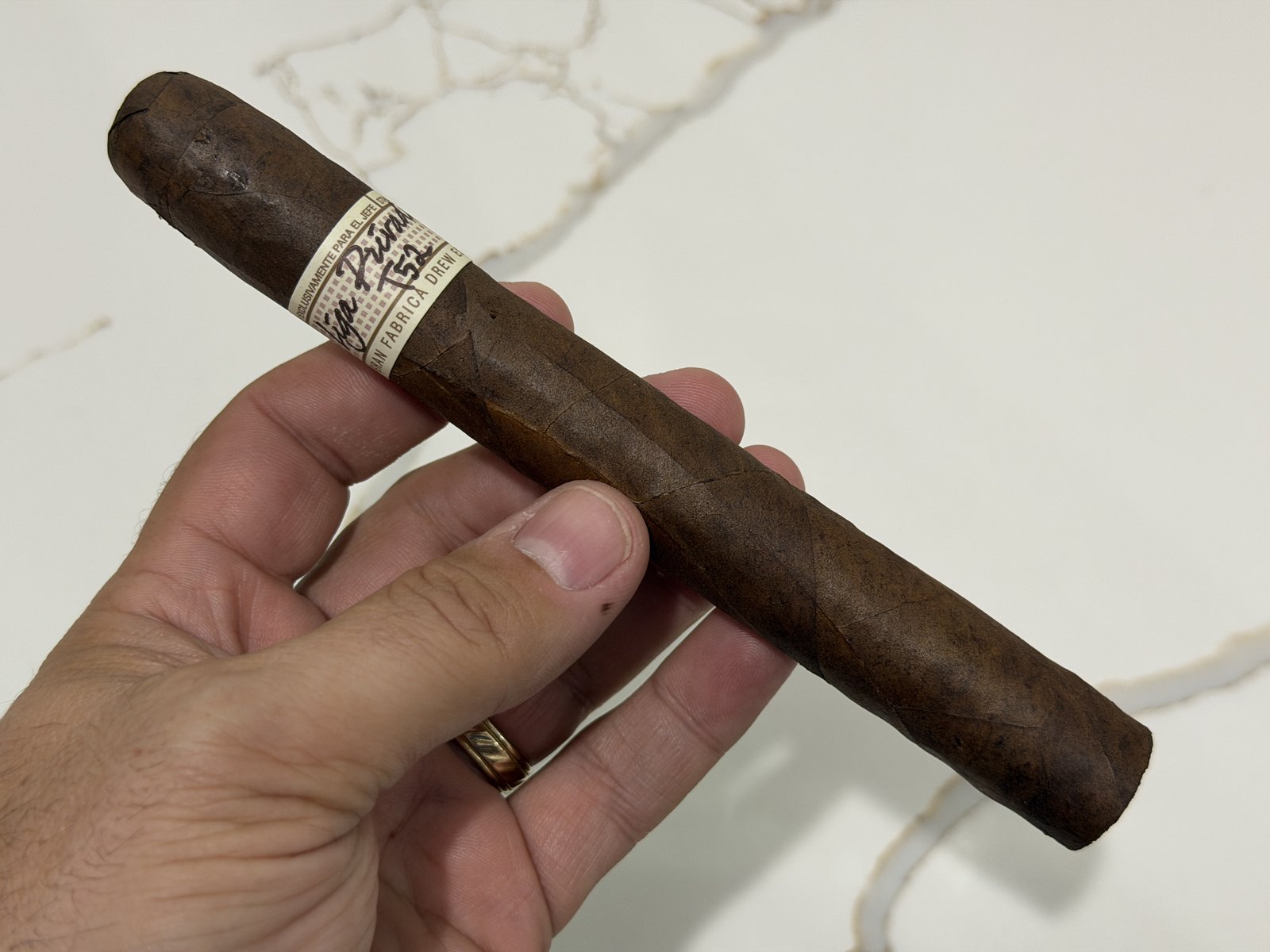 Unlit Liga Privada T52 Corona Doble held over a white marble counter, medium-dark brown wrapper with a double cap and the cream-and-gold Liga Privada T52 band