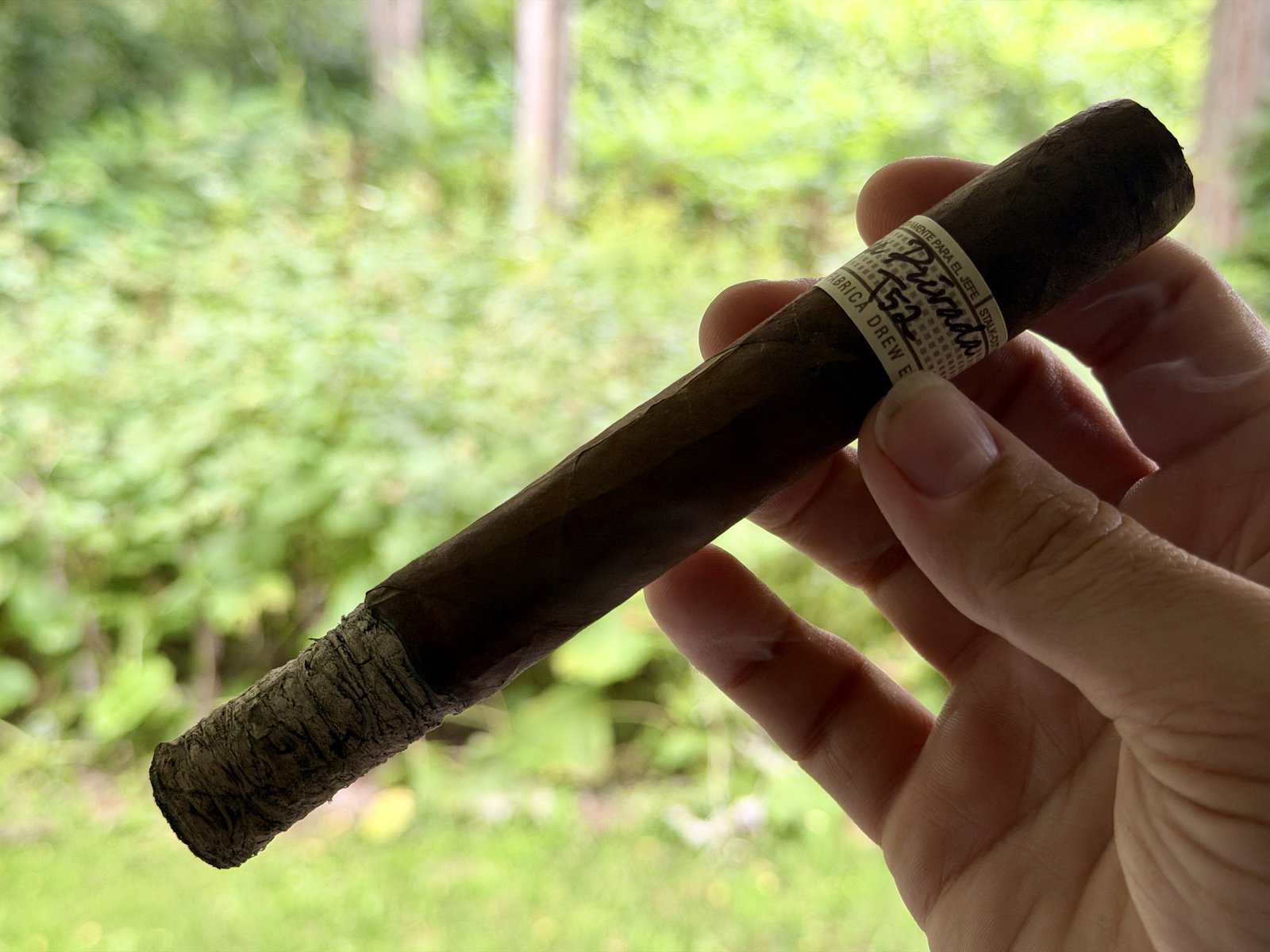 Lit Liga Privada T52 Corona Doble held outdoors with green foliage behind it, band on, foot just starting to ash