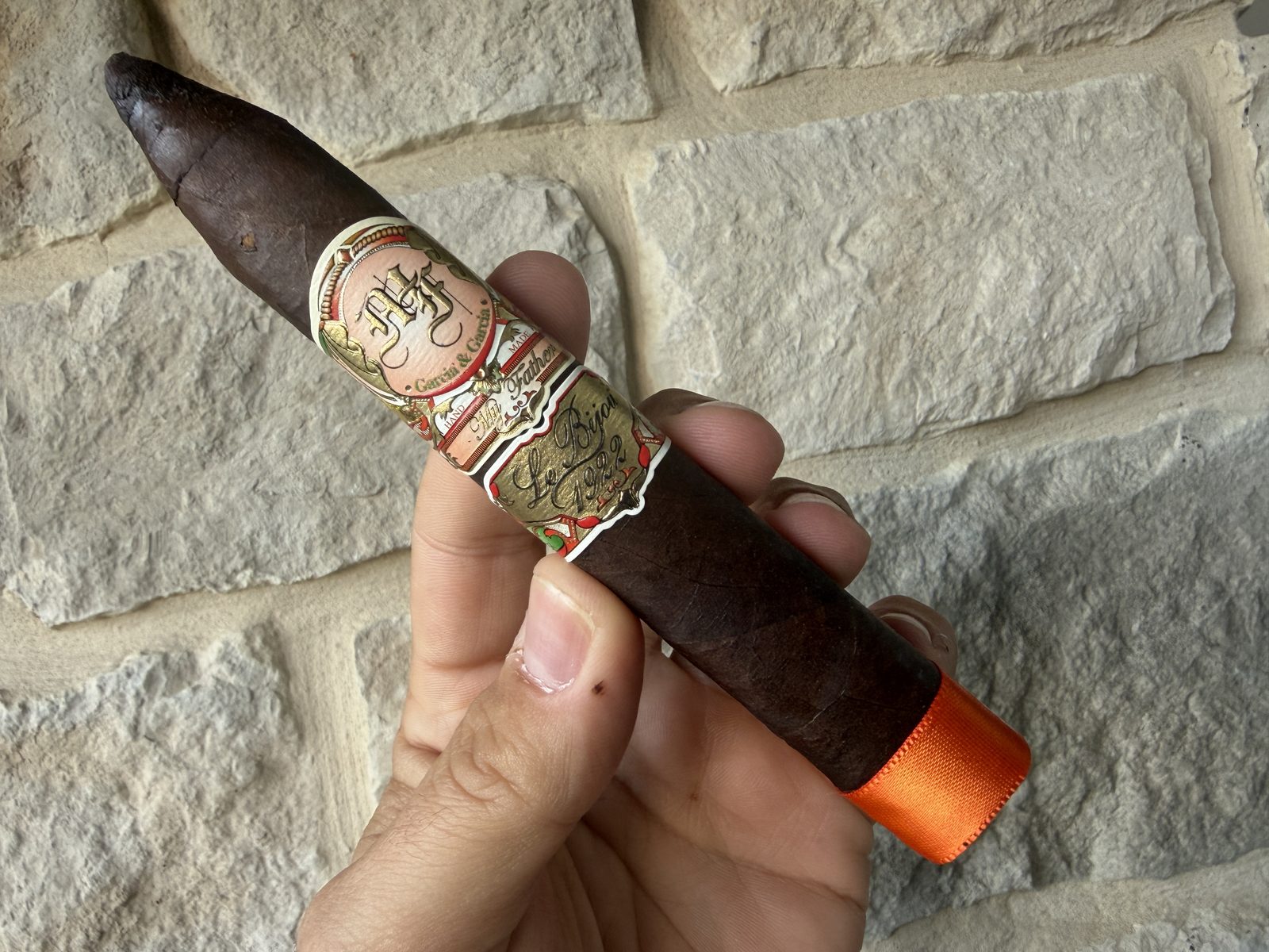 My Father Le Bijou 1922 Torpedo held in hand before lighting, dark oily wrapper, ornate bands and bright orange foot ribbon