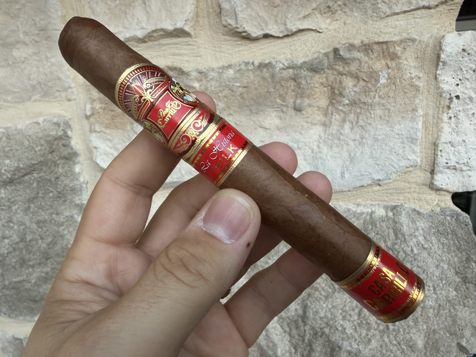 Unlit Perez-Carrillo La Historia Silk Toro held in hand against a stone wall, light brown wrapper with red and gold main band, secondary La Historia Silk band, and Casa Carrillo foot band