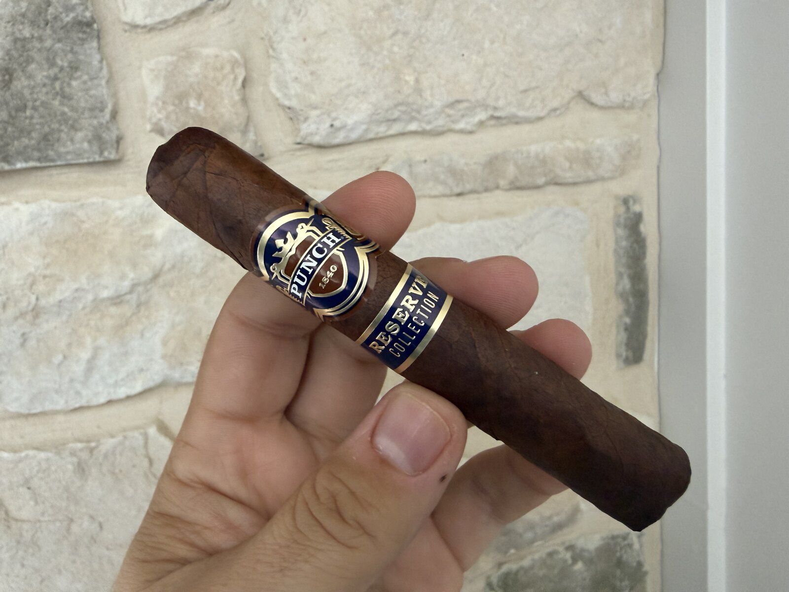 Punch Reserve Collection Robusto held in hand against a light stone wall, dark espresso-brown wrapper with a navy and gold Punch 1840 band and a Reserve Collection secondary band