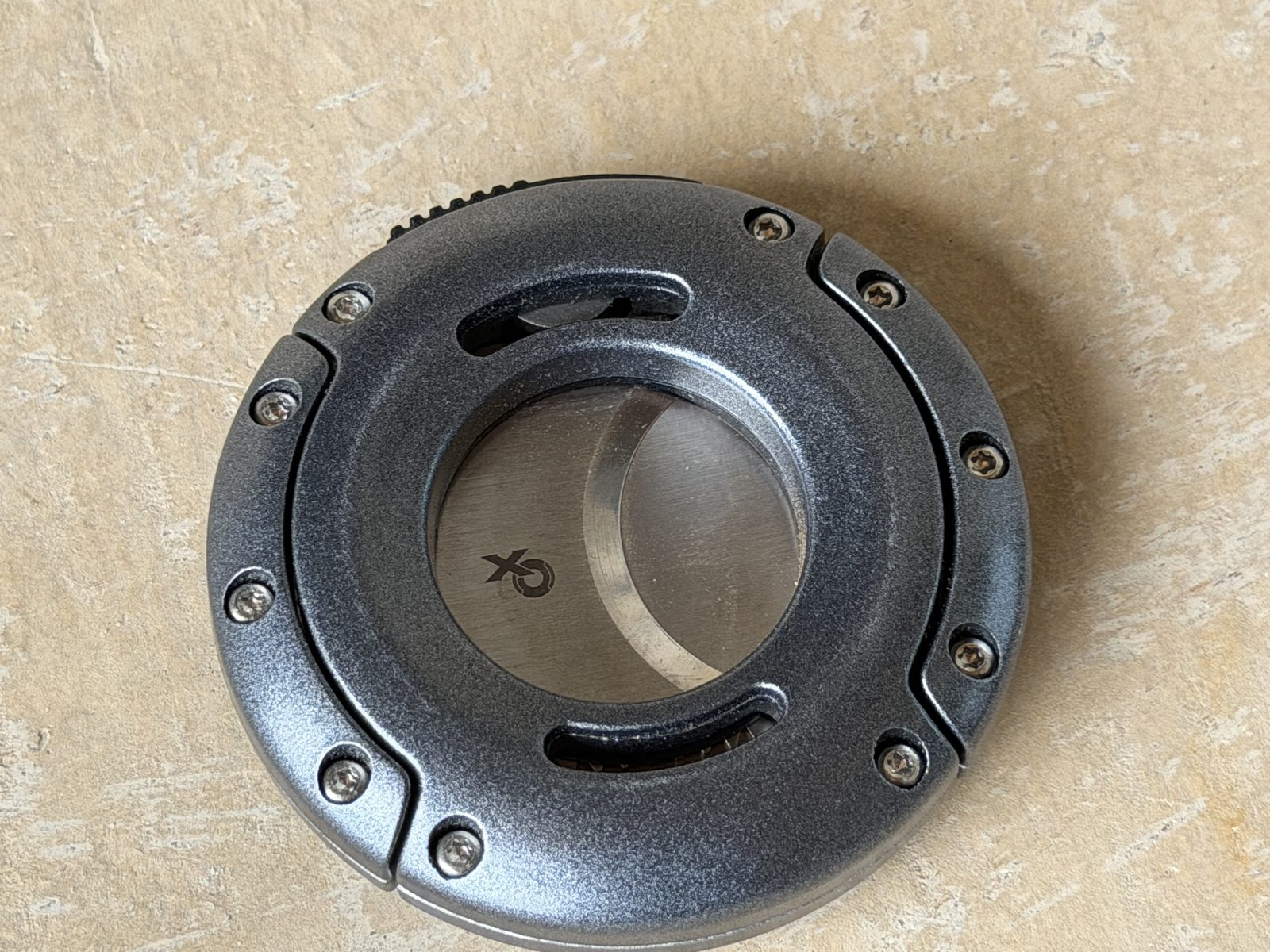 Xikar XO cutter in gunmetal, closed, round body with exposed screws on a stone surface